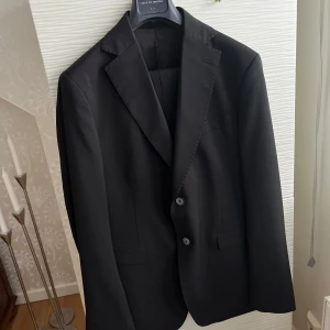 Svart kostym från Sir of Sweden - Elegant svart kostym från Sir of Sweden. Kavajen har klassisk design med två knappar och byxorna har en stilren passform.                                 Modellen heter Sven, storlek 52L. Ett litet lagat hål finns på byxorna enligt bild. Så vi kan säja att byxorna gånger med på köpet.            Beninnerlängd 87 cm, midja 90 cm, ärm 68 cm, längd axel och ner 75 cm.
