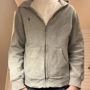 Grå hoodie från Ralph Lauren - Säljer en stilren grå hoodie från Ralph Lauren med dragkedja och klassisk logga på bröstet.    På mig är den liten då jag är 171cm, så jag skulle säga att den passar en runt 160cm. Priset är inte hugget i sten. Har du frågor eller funderingar är de bara att ställa.