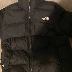 Svart dunjacka från The North Face - Säljer en svart dunjacka från The North Face med dragkedja och hög krage. Perfekt för kalla dagar med sin pufferdesign som håller värmen. Jackan har en klassisk look med logotyp på bröstet.