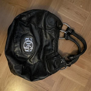 Svart axelväska med logotyp - Snygg svart axelväska från Guess med präglat mönster och en stor logotyp i silver på framsidan. Väskan har dubbla handtag och metallringar som detaljer. Stor och rymlig - rymmer ex en laptop. Hänger väldigt snyggt på axeln och ger en cool y2k-look. 💗
