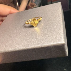 Guldfärgad ring med blomdetalj - 21k van cleef guldring storlek 18. 1,5gram Elegant guldfärgad ring med en vacker blomformad detalj i mitten. Ringen har en tunn, vriden design som ger en unik och stilren look. Perfekt för att lägga till en touch av elegans till din outfit.