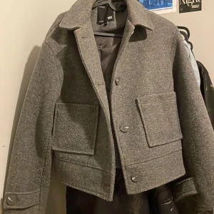 Grå kappa från 157 - Snygg grå kappa från 157 i en croppad stil med stora fickor och knappar framtill. Perfekt för höst och vår med sin stilrena design och bekväma passform.