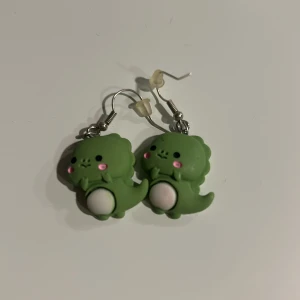 Gröna dinosaurieörhängen - Söta gröna örhängen formade som små dinosaurier med rosa kinder och vita magar. Perfekta för att ge en lekfull touch till din stil. De är lätta och bekväma att bära hela dagen.Tvättar innan skickar.