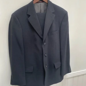 Mörkgrå kostym från Oscar Jacobson - Säljer en stilren mörkgrå kostym från Oscar Jacobson. Kavajen har en klassisk design med tre knappar och fickor med lock. Insidan är fodrad med ett elegant grått tyg och har märkesdetaljer. Byxorna matchar kavajen perfekt och har en rak passform. Perfekt för både formella tillfällen och fest.