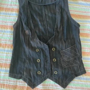 Svart randig väst från Vila - Säljer en snygg svart/brun väst från Vila i storlek L. Västen har ett randigt mönster och är försedd med knappar framtill samt en dekorativ ficka. Den har också ett justerbart spänne på baksidan för bättre passform. Perfekt för en stilren och klassisk look!