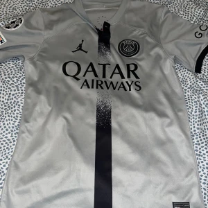 Grå PSG fotbollströja med Hakimi #2 - Säljer en grå PSG fotbollströja från Jordan med Hakimi #2 på ryggen. Tröjan har korta ärmar och är i nyskick. Den har Qatar Airways som sponsor på framsidan och GOAT på ärmen. Perfekt för alla PSG-fans!
