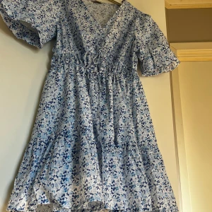 Blommig sommarklänning med volanger - Säljer en superfin blommig klänning i ljusblått och vitt. Den har en v-ringad hals och korta ärmar med volanger. Klänningen är perfekt för sommaren med sitt lätta och luftiga material. Den har en knytning i midjan för en smickrande passform och volanger längst ner för extra stilpoäng. Perfekt för en dag på stan eller en sommarfest! Aldrig använd. nypris: 500kr