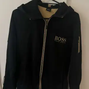 Säljer en snygg svart hoodie från Hugo Boss. Den har en dragkedja framtill och en huva för extra komfort. Perfekt för en casual look eller att mysa i hemma. Boss-loggan är tryckt på bröstet för en stilren touch.