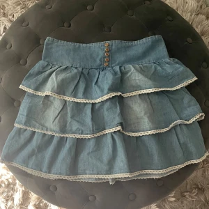 Söt jeanskjol med volanger och spetsdetaljer - Säljer en superfin jeanskjol med volanger och söta spetsdetaljer😍😍 super fina detaljer på hela kjolen, knappar spets osv❤️‍🔥❤️‍🔥