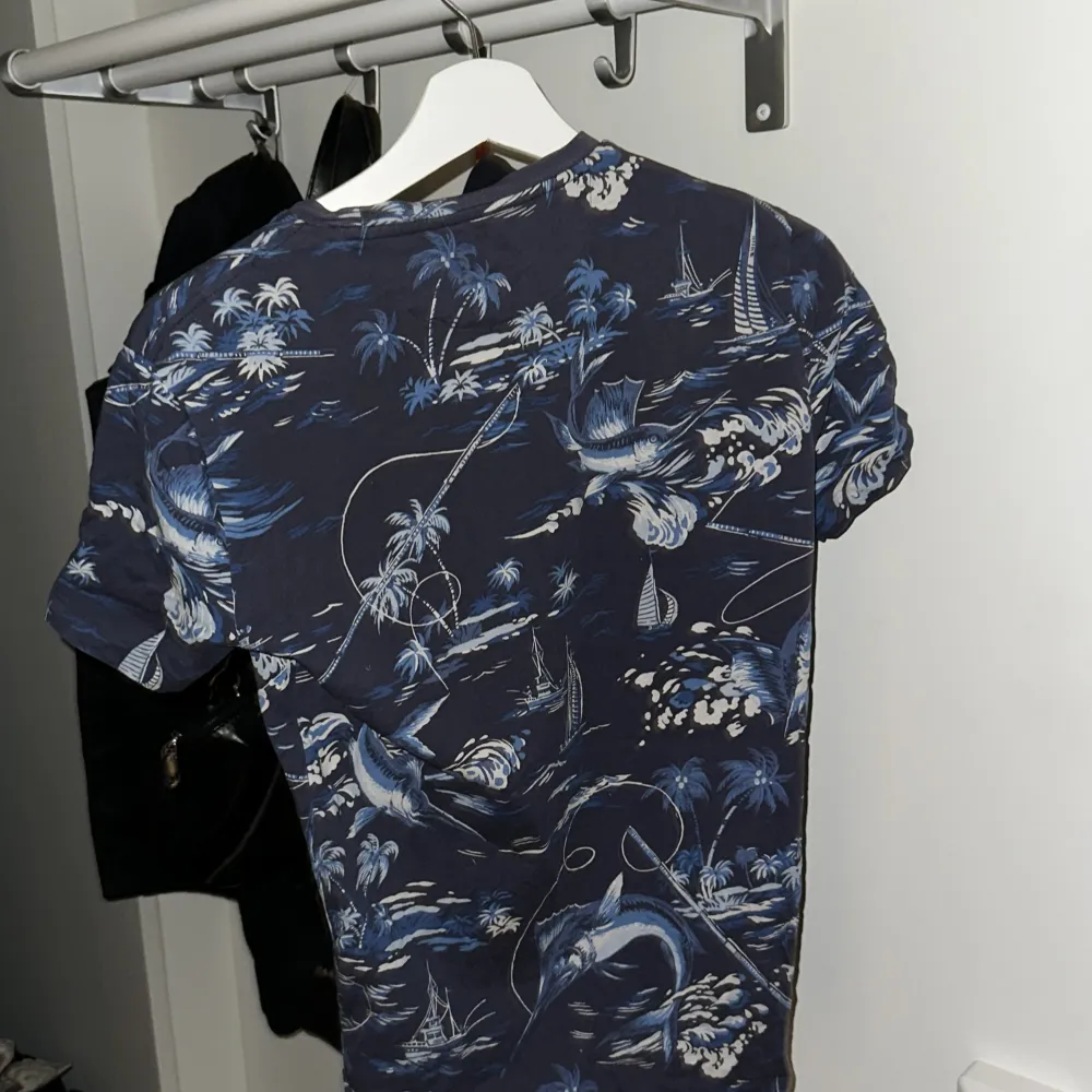 Säljer en snygg blå t-shirt från Polo Ralph Lauren med ett coolt mönster av palmer och båtar. T-shirten är kortärmad och har en slim passform, perfekt för sommarens äventyr. Materialet känns mjukt och skönt mot huden. Passar både till vardags och fest!. T-shirts.