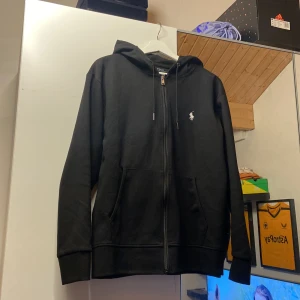 Polo Ralph Lauren - Säljer en stilren svart hoodie från Ralph Lauren med dragkedja och huva. Den har en broderad logga på bröstet och är tillverkad i mjukt material. Perfekt för en avslappnad look.