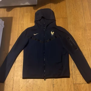 Svart huvjacka från Nike - Säljer en blå huvjacka från Nike med dragkedja och huva. Tröjan har en logotyp på bröstet och är långärmad. Perfekt för en sportig look eller avslappnade dagar.