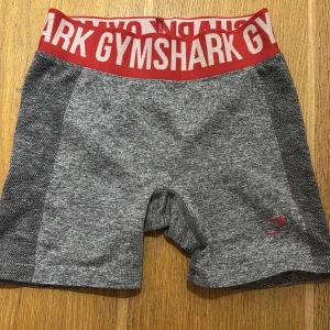 Grå träningsshorts från Gymshark - Snygga grå träningsshorts från Gymshark med röd resår i midjan och logga. Perfekta för gymmet eller löprundan. 