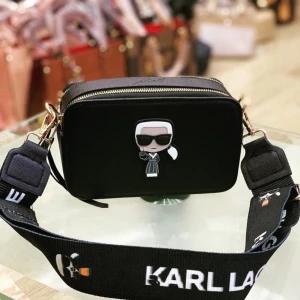 Svart axelväska från Karl Lagerfeld - Snygg svart axelväska med en cool Karl Lagerfeld-figur på framsidan. Väskan har en justerbar axelrem med logotyp och en praktisk dragkedja upptill. Perfekt för att ge din outfit en stilfull touch!
