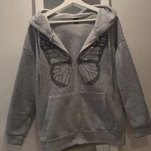 Grå hoodie med fjärilsdetalj - Säljer en grå hoodie med dragkedja och en stor fjärilsdetalj i glittrande stenar på framsidan. Tröjan har en bekväm passform med långa ärmar och en praktisk huva. Perfekt för en avslappnad stil med en touch av glamour.