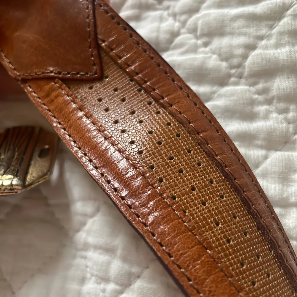 Snyggt brunt skärp i skinn med ett unikt guldfärgat spänne. Perfekt för att ge en stilfull touch till din outfit. Skärpet har en längd på 75-100 cm.. Asusteet.
