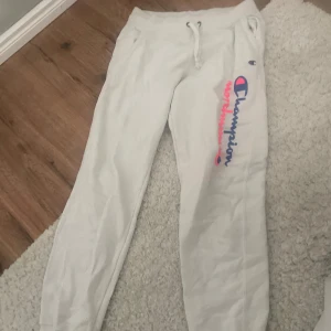 Vita mjukisbyxor från Champion - Säljer ett par vita mjukisbyxor från Champion med rosa och blå logga längs benet. Byxorna har snörning i midjan och är perfekta för en avslappnad stil.