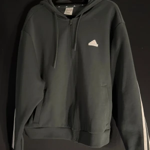 Grå hoodie från Adidas - Säljer en grå hoodie från Adidas med dragkedja och klassiska vita ränder längs ärmarna. Tröjan har en justerbar huva och två fickor framtill. Perfekt för en sportig look eller avslappnad stil.