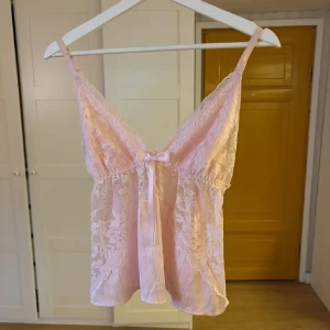 Rosa spetslinne med rosett - Säljer ett sött rosa linne med spetsdetaljer och en liten rosett framtill. Linnet har smala axelband och en vacker spetsdesign som ger en romantisk känsla. Perfekt för varma sommardagar eller som ett fint lager under en kofta. Passar även i S🩷