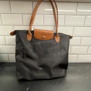 Svart axelväska från Longchamp - Snygg svart väska som LIKNAR en från Longchamp med bruna skinnhandtag och en praktisk dragkedja. Väskan har en klassisk design med en knappdetalj framtill och är perfekt för att bära med sig det nödvändigaste. En stilren accessoar för alla tillfällen.