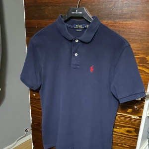 Mörkblå pikétröja från Ralph Lauren - Säljer en klassisk mörkblå pikétröja från Ralph Lauren med den ikoniska röda loggan på bröstet. Tröjan har korta ärmar och en traditionell krage med knappar. Perfekt för en stilren och avslappnad look.