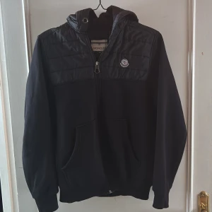 Svart jacka med huva från Moncler - Snygg svart jacka från Moncler med huva och dragkedja. Jackan har ett stilrent märke på bröstet och är perfekt för kyligare dagar. Den har långa ärmar och en quiltad design upptill.