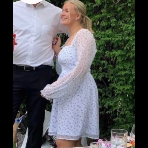 Vit blommig klänning - Charmig vit klänning med blå blommor och långa puffärmar. Den har en fyrkantig urringning och en kort längd som ger en söt och somrig look. Perfekt för en solig dag i trädgården eller en sommarfest.