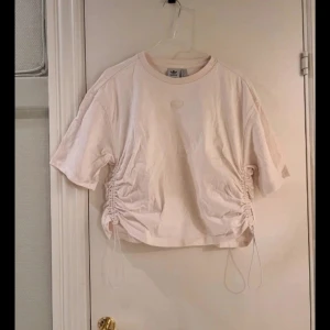 Beige kortärmad topp från Adidas - Säljer en beige kortärmad topp från Adidas med snörning på sidorna för en justerbar passform. Toppen har en rund halsringning och Adidas-loggan broderad på bröstet. Perfekt för en avslappnad stil.