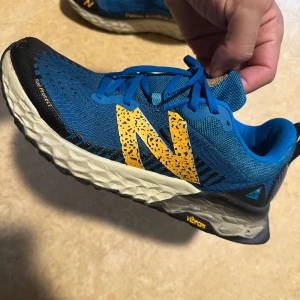 Blå trail-skor från New Balance - Säljer ett par blå trail-skor från New Balance med gul logga och svarta detaljer. Skorna har snörning och en robust Vibram-sula för bra grepp. Perfekta för terränglöpning, gym mm. Skorna är använda ett fåtal gånger, legat ståendes mestadels, syns på undersidan. Lägger ut skorna biligt, inga prissänkningar här.