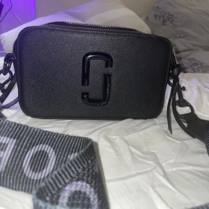 Svart axelväska från Marc Jacobs - Snygg svart axelväska från Marc Jacobs med ett stilrent design. Väskan har en justerbar axelrem med märkets logga och två dragkedjefack för enkel organisering. Perfekt för att bära dina nödvändigheter med stil.