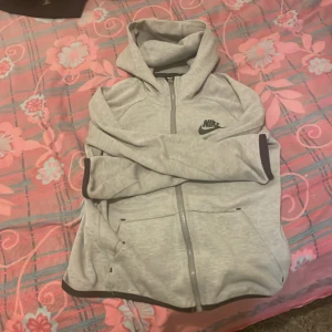 Grå hoodie från Nike - Säljer en grå hoodie från Nike med dragkedja och fickor framtill. Den har en klassisk Nike-logga på bröstet och är perfekt för en avslappnad stil. Muddar i ärmslut och nederkant ger en skön passform. Obs! Den är L i barnstorlek. Kan t.om gå ner i pris lite