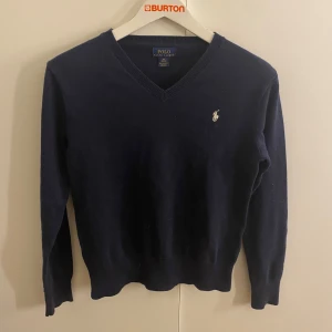 Mörkblå tröja från Polo Ralph Lauren - Säljer en stilren mörkblå tröja från Polo Ralph Lauren med v-ringning och det klassiska logotypbroderiet på bröstet. Tröjan har långa ärmar och är perfekt för en elegant vardagslook.