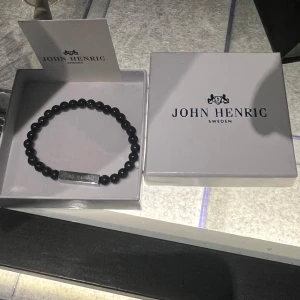 Svart armband från John Henric - Elegant svart armband från John Henric med runda pärlor och en metallbricka med ingraverad logotyp. Perfekt accessoar för en stilren look. Levereras i originalförpackning.
