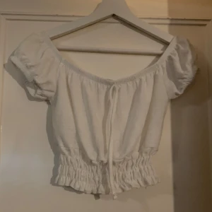 Vit kortärmad topp - Säljer en söt vit topp med korta puffärmar och elastisk midja. Toppen har en knytning framtill och är croppad, vilket ger en trendig look. Perfekt för varma dagar!