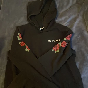 Svart hoodie med rosor från Gina Tricot - Säljer en svart hoodie från Gina Tricot med texten 'NO THANKS.' på bröstet. Ärmarna är dekorerade med röda rosor och gröna blad. Perfekt för en avslappnad stil. 🖤🌹