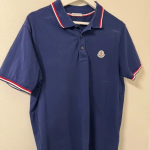 Blå pikétröja från Moncler - Snygg blå pikétröja från Moncler med korta ärmar och klassisk krage. Tröjan har röda och vita detaljer på kragen och ärmsluten samt Monclers logotyp broderad på bröstet. Perfekt för en stilren look.