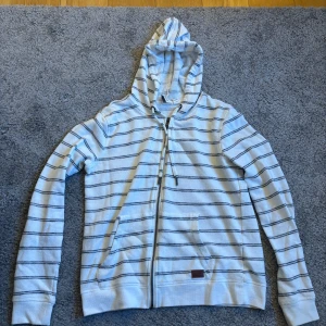 Vit och svart randig hoodie från Roxy - Säljer en vit och svart randig hoodie från Roxy. Den har en dragkedja framtill och en praktisk huva med snören. Perfekt för en avslappnad stil.