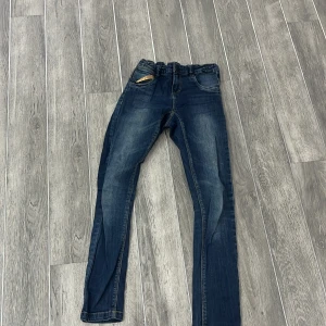 Blå jeans med paljettdetaljer från Name It - Snygga blå jeans från Name It med glittriga paljettdetaljer på bakfickorna. Perfekta för att ge lite extra stil till vardagslooken. De har en bekväm passform och är tillverkade i ett mjukt denimtyg.