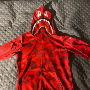 Röd camo hoodie med hajmotiv - Säljer en cool röd camo hoodie från A Bathing Ape med ett hajmotiv på huvan. Hoodien har en dragkedja framtill och är långärmad. Perfekt för dig som vill sticka ut med en unik stil!