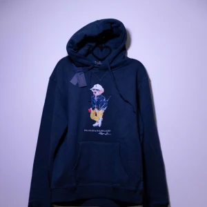 Mörkblå hoodie från Polo Ralph Lauren - Säljer en helt ny hoodie från Polo Ralph Lauren med en cool broderad nalle på framsidan. Hoodien har en klassisk känguruficka och justerbar huva med dragsko. Perfekt för en avslappnad stil.