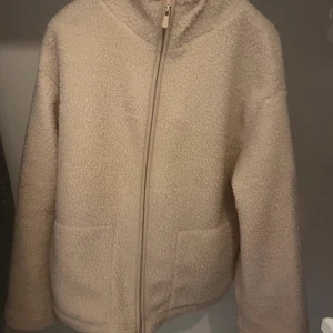 Beige teddyjacka med dragkedja - Mysig beige teddyjacka med dragkedja framtill. Jackan har en enkel design med två fickor och är perfekt för kyligare dagar. Den har långa ärmar och en mjuk, fluffig yta som ger en avslappnad look.