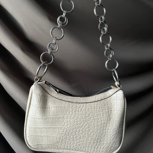 Beige axelväska med kedjedetalj - Snygg beige axelväska med krokodilpräglat mönster och en cool kedja som axelrem. Väskan har en dragkedja upptill för säker förvaring. Perfekt för att ge din outfit en trendig touch.