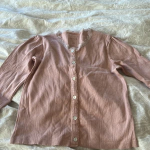 Rosa kofta med spetsdetaljer st 128 - Söt rosa kofta  med spetsdetaljer runt halsringningen. Den har knappar framtill och långa ärmar. Perfekt för en romantisk look eller en mysig dag.