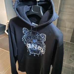 Svart hoodie från Kenzo - Säljer en svart hoodie från Kenzo med det ikoniska tigerhuvudet i blått på framsidan. Tröjan har en klassisk passform med huva och dragsko. Perfekt för en stilren och bekväm look.