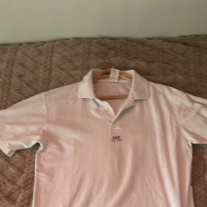 Vit pikétröja från Helly Hansen - Säljer en klassisk vit pikétröja från Helly Hansen. Tröjan har en krage med knappar och det ikoniska HH-logotypen på bröstet. Perfekt för en stilren och avslappnad look.