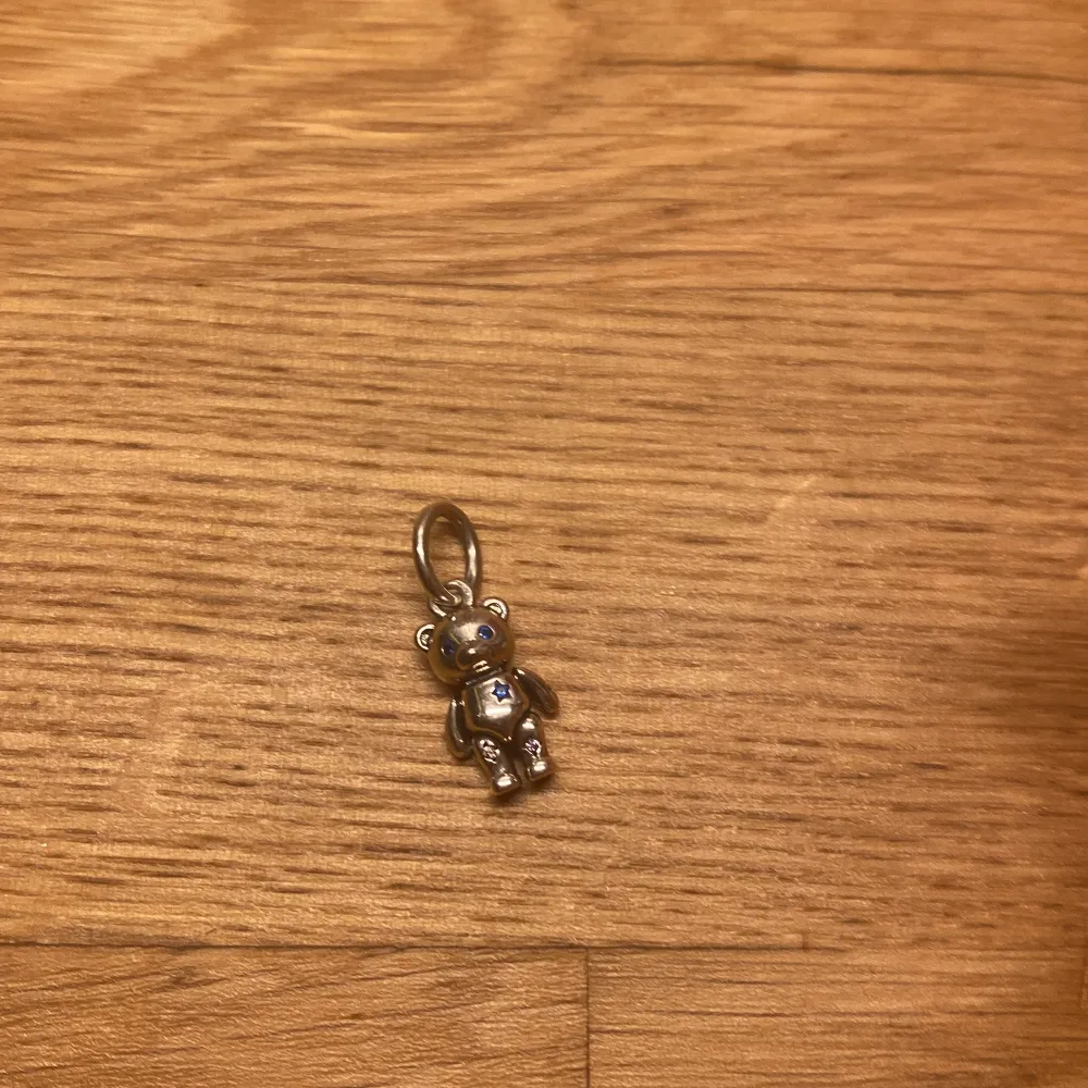 En liten nalle berlock från Pandora i silver med små stenar i färgen blå. Nallens armar och ben kan röra på sig. . Asusteet.