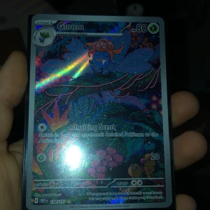 Gloom Pokémonkort - Säljer ett holografiskt Gloom Pokémonkort med 80 HP. Kortet har en färgglad och glittrande design med Gloom i en mystisk skogsmiljö. Perfekt för samlare eller Pokémon-entusiaster! Den har lite vit på en kant