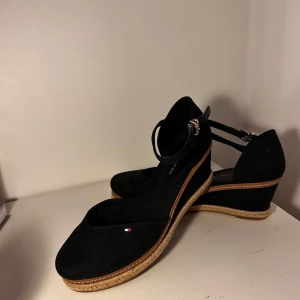 Mörk blå kilklackar från Tommy Hilfiger - Snygga mörkblå kilklackar från Tommy Hilfiger med en elegant remdesign och spänne. Skorna har en bekväm kilklack och en liten logodetalj på sidan. Perfekta för att ge lite extra höjd och stil.