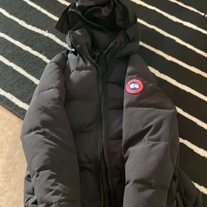 Svart dunjacka från Canada Goose - Säljer en svart dunjacka från Canada Goose med huva och dragkedja. Jackan har en klassisk design med två framfickor och en broderad logotyp på bröstet. Perfekt för kalla dagar.