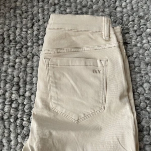 Beige byxor från IVY - Snygga beige byxor från IVY med hög midja och raka ben. Byxorna har en klassisk design med fem fickor och en dragkedja framtill. Perfekta för en stilren look. Storlek 27/28.